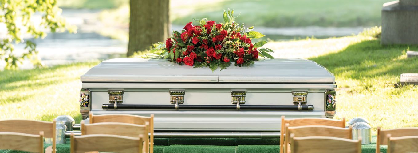 A white coffin with red flowers, surrounded by chairs, on green grass. Outdoor setting.