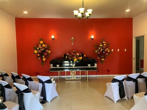 Funeral service room with black casket, floral arrangements, and seated guests. Red wall backdrop.