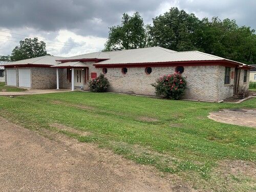 Beige-stoned ranch house with red trim, circular windows, and a metal roof. Green lawn and overcast sky.