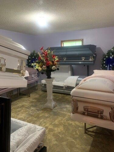 Funeral home interior with several closed caskets, floral arrangements, and seating.