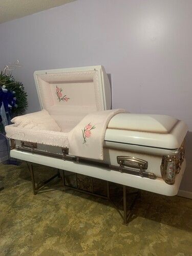 White open casket with pink interior and floral embroidery on a gold stand.
