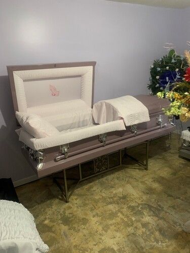 Lavender casket with white interior, open, on silver stand in a room, flowers in the background.
