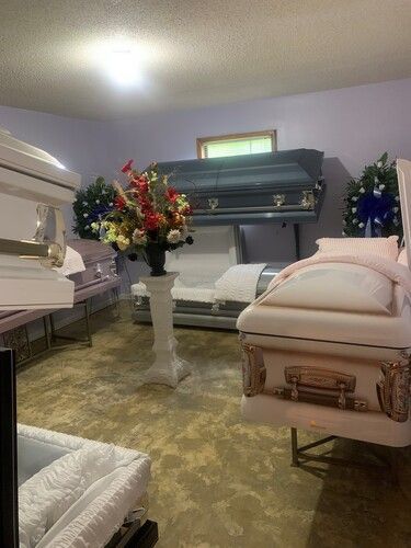 A funeral home showroom displaying various caskets, with floral arrangements and neutral-toned walls.