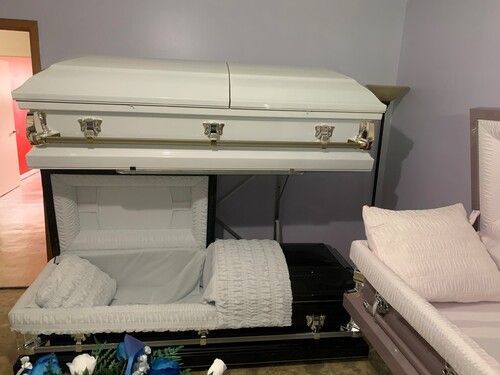 Caskets displayed in a funeral home. One is open with pillows. One is closed above. Another is partially visible on the right.