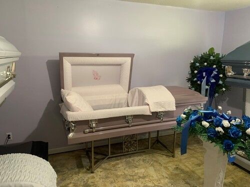 A lilac casket on a stand, open with white interior, in a room with floral arrangements.