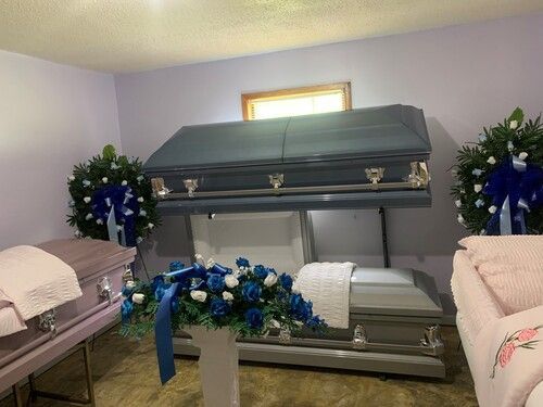 Funeral home interior with open and closed caskets, floral arrangements, and light blue walls.