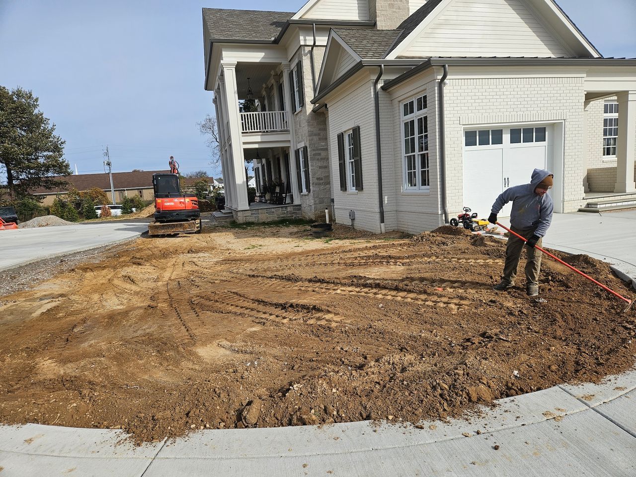 Man raking dirt near a house under construction; small excavator visible.
