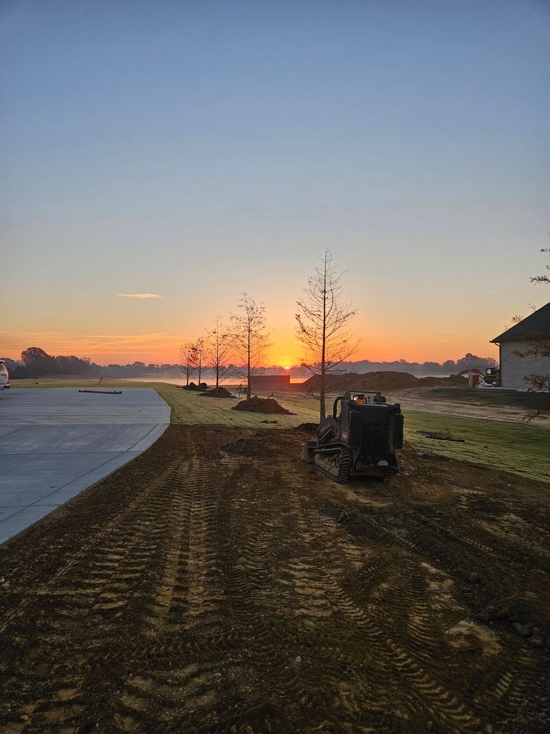 Sunrise over construction site with trees, dirt, a machine, and a road.