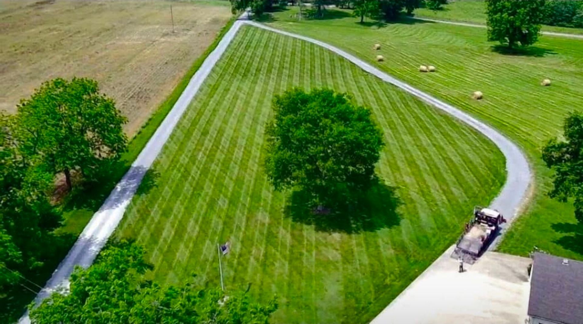 Aerial view of a long driveway with mowed lawn and hay bales, a tree in the center, and a truck.