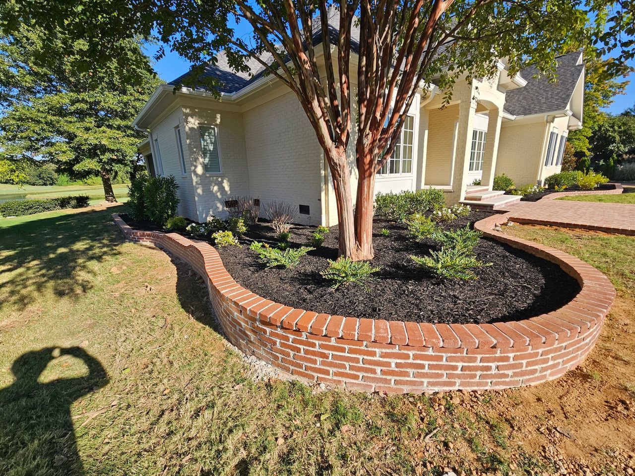 Brick-edged flower bed with mulch, tree, and plants in front of a light yellow house.