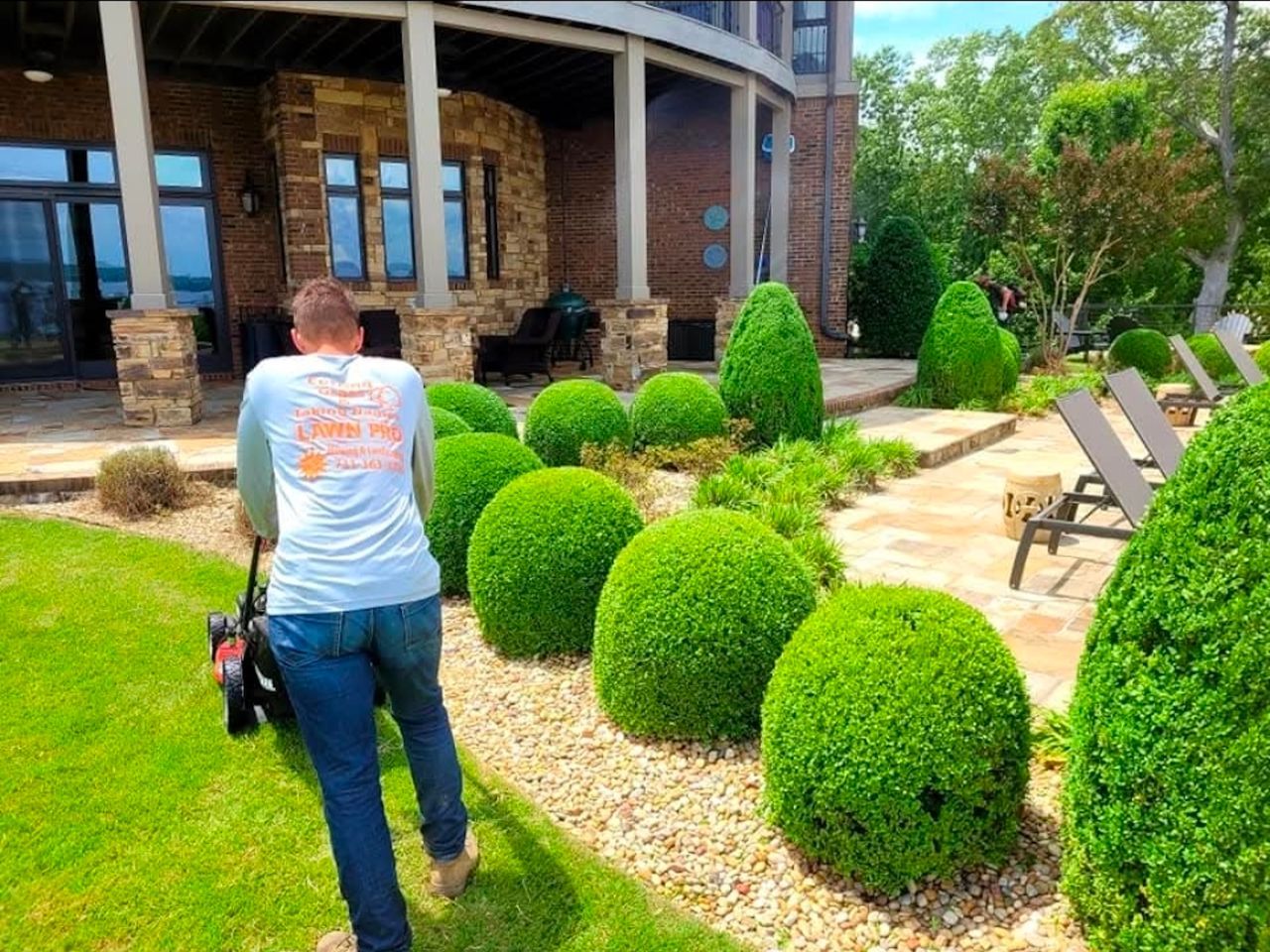 Man mowing lawn next to well-manicured yard with bushes and house.