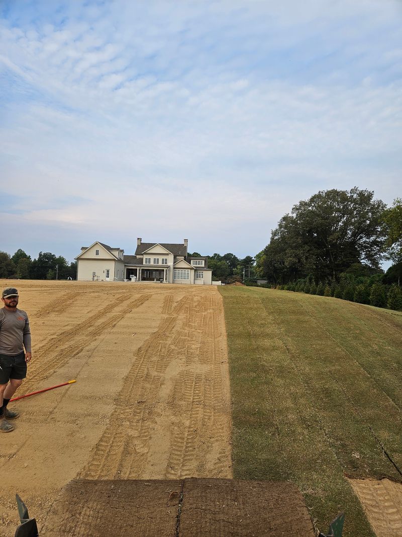 Man standing on soil next to freshly sodded lawn with a house in the background. Cloudy sky.
