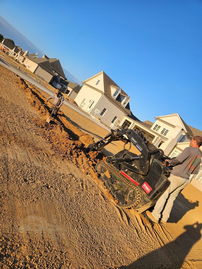 A person operating an excavator on a construction site with houses in the background under a blue sky.