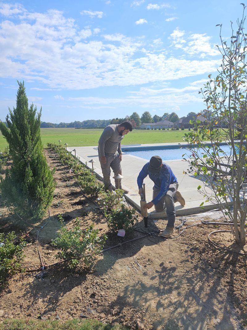 Two people landscaping near a pool, sunny day, blue sky, field in background.
