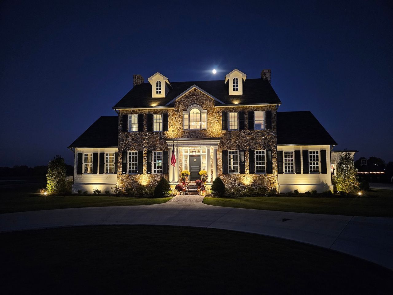 Large stone house lit at night, driveway leads to the front door, blue sky with stars above.