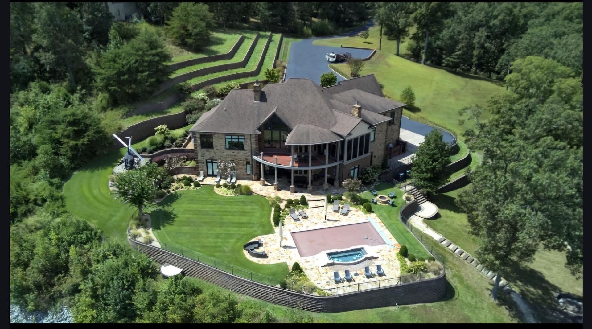 Large house with pool on a grassy hill, surrounded by trees and a retaining wall.