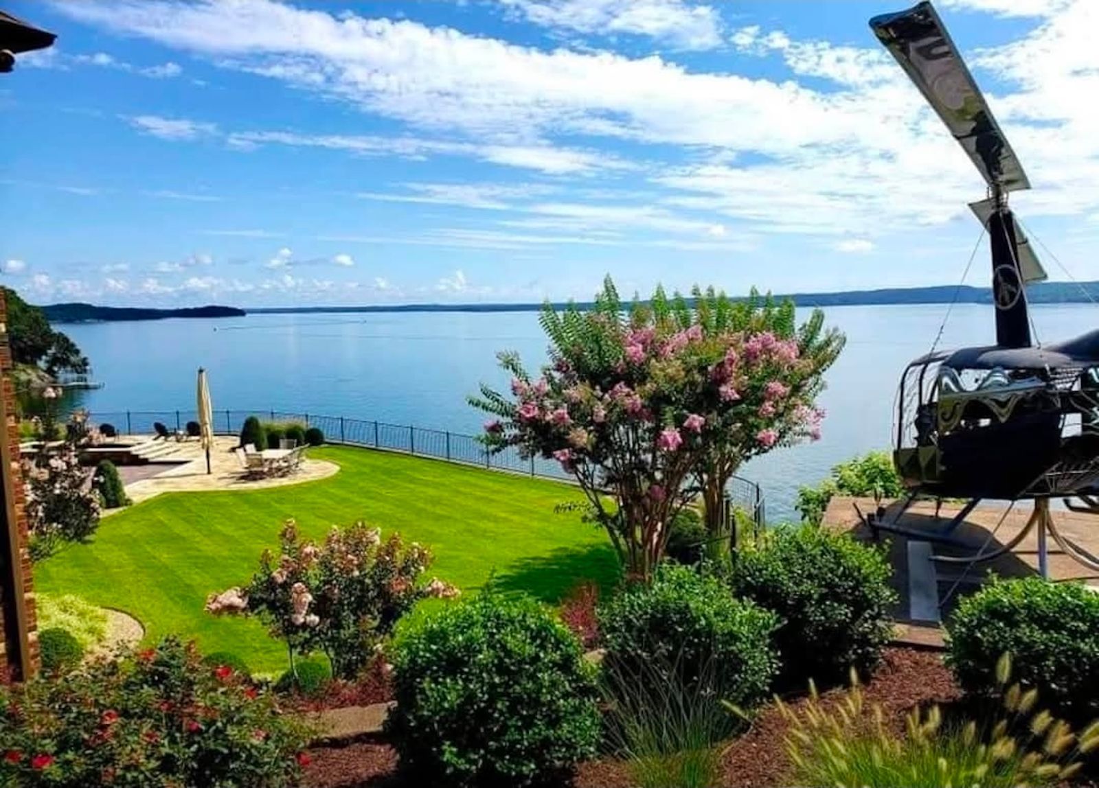 Lush green lawn with lake view, pink flowering tree, helicopter, and sunny blue sky.