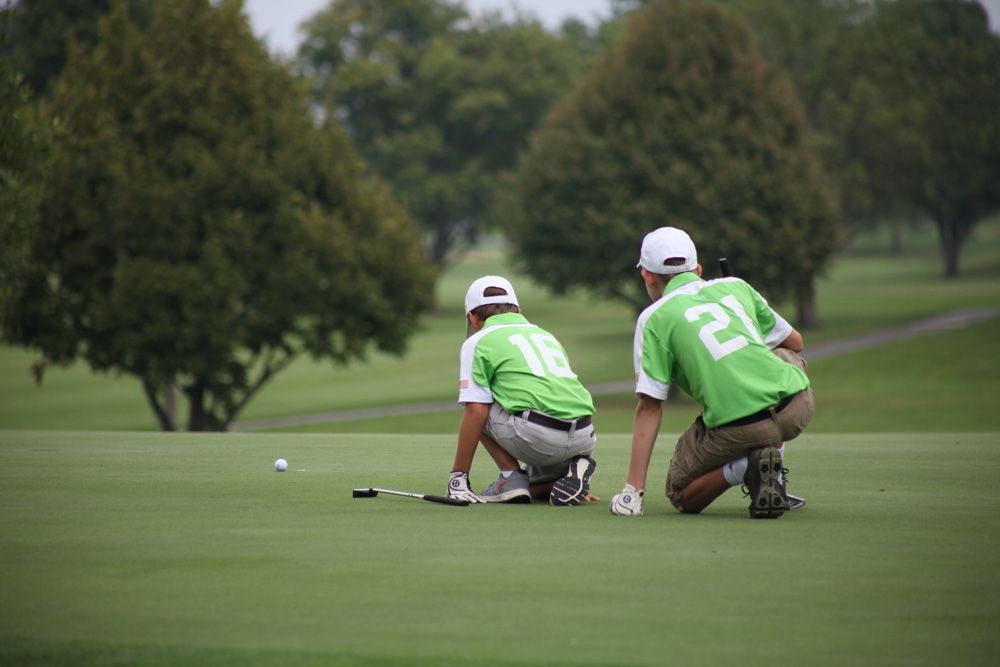 Two golfers in green shirts, kneeling on a green putting green, assessing a golf ball near the hole.
