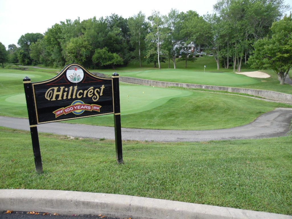 Hillcrest Golf Club sign with putting green in background. Green grass, trees, and a curved path.