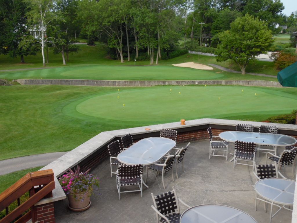 Patio overlooking a golf course green with tables and chairs, green grass, and trees in the background.