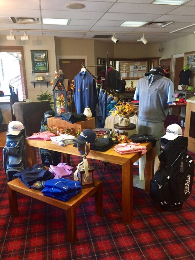 Golf shop interior with clothing displays on wood tables, golf bags, and mannequins. Red plaid carpet.