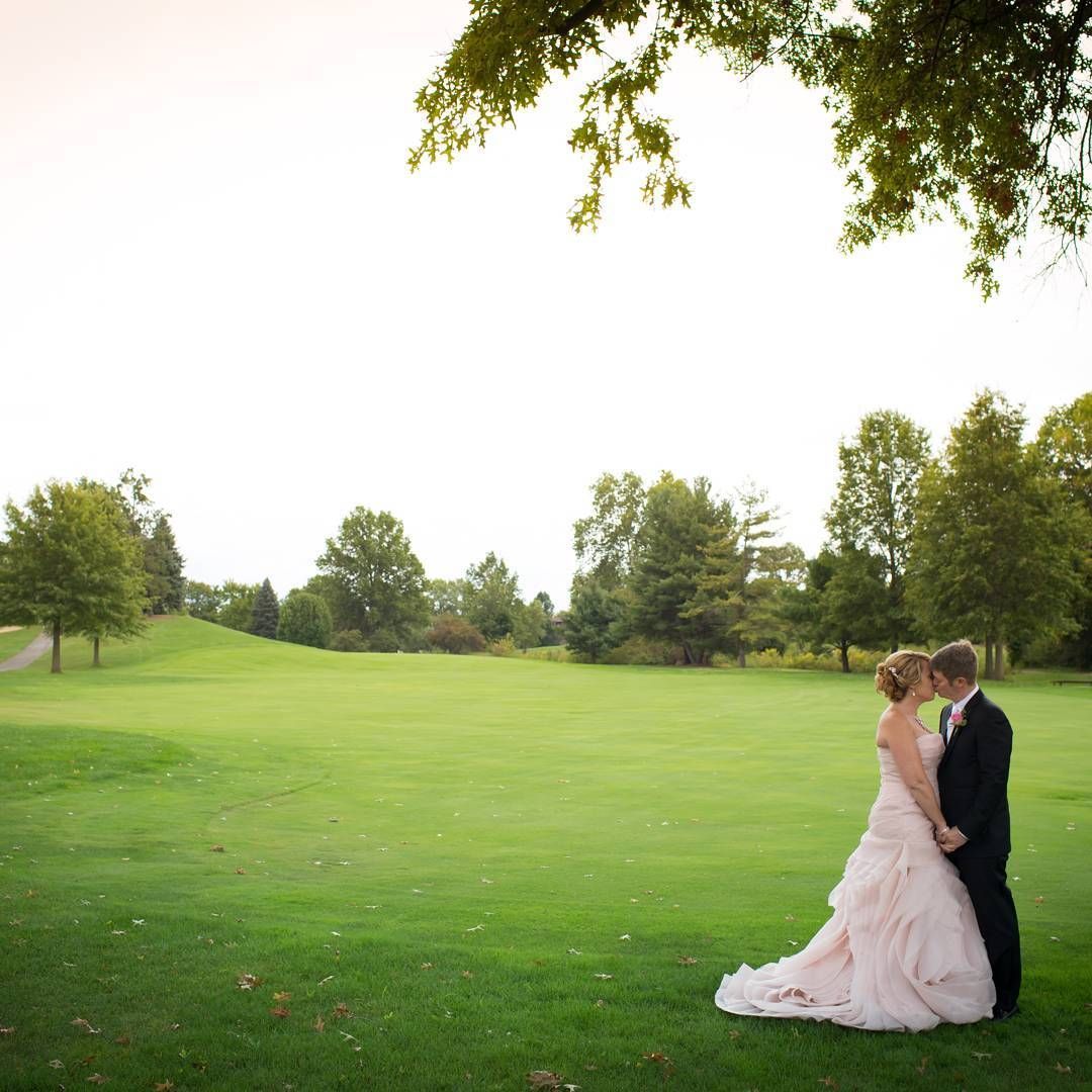 Bride and groom kiss on a green golf course; she wears a pink dress, he wears a black suit.