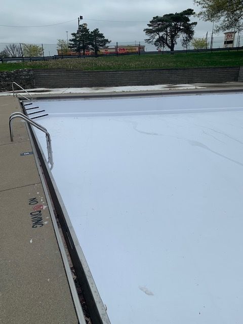 Empty outdoor swimming pool, covered with a white tarp. Metal ladder on the side. "No Diving" painted.