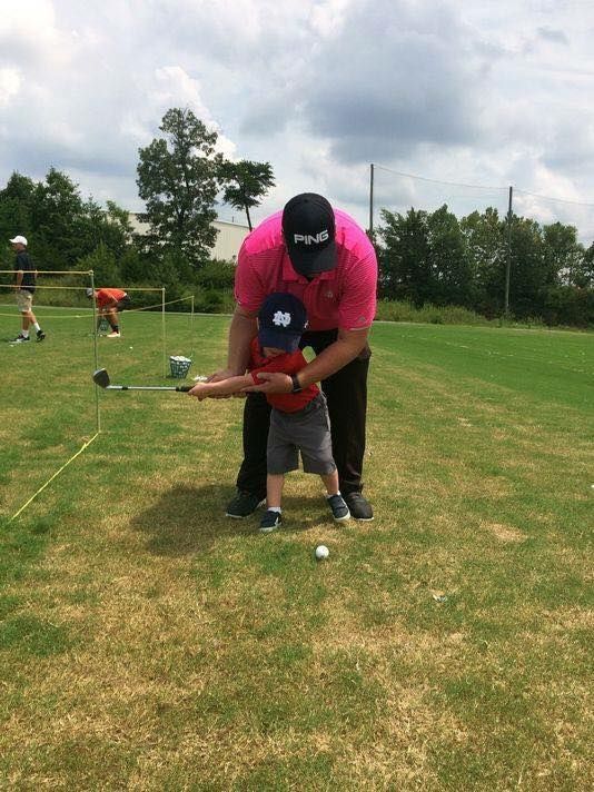 Man in pink shirt helps toddler swing a golf club on a green course.