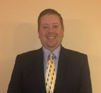 Man in a suit and tie, smiling at the camera against a cream-colored background.