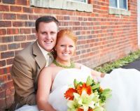 Bride and groom smiling together in front of a brick wall; bride holds a colorful bouquet.