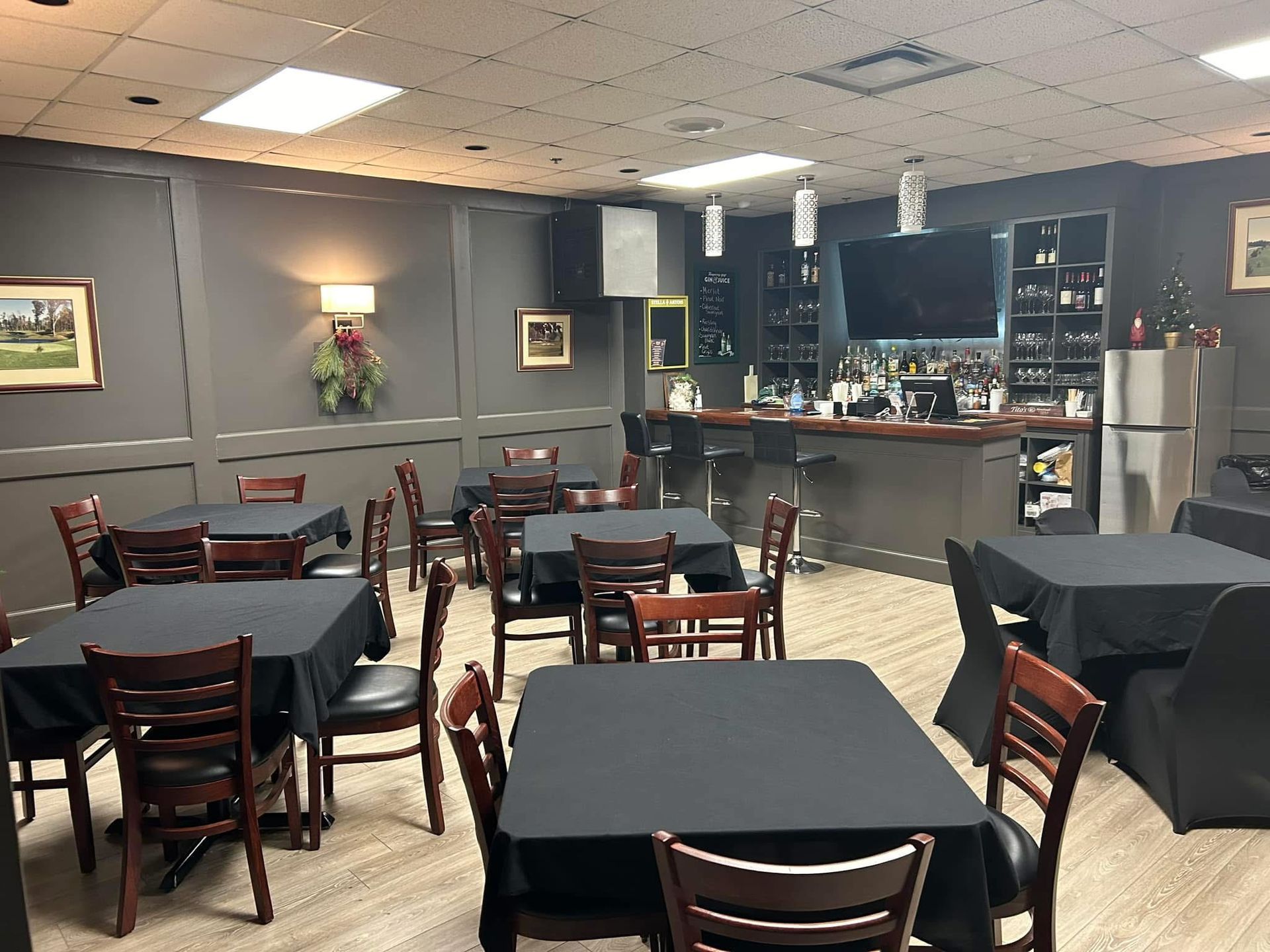 Restaurant with tables, bar, and stools. Tables have black tablecloths, chairs are wooden. Gray walls and lighting.