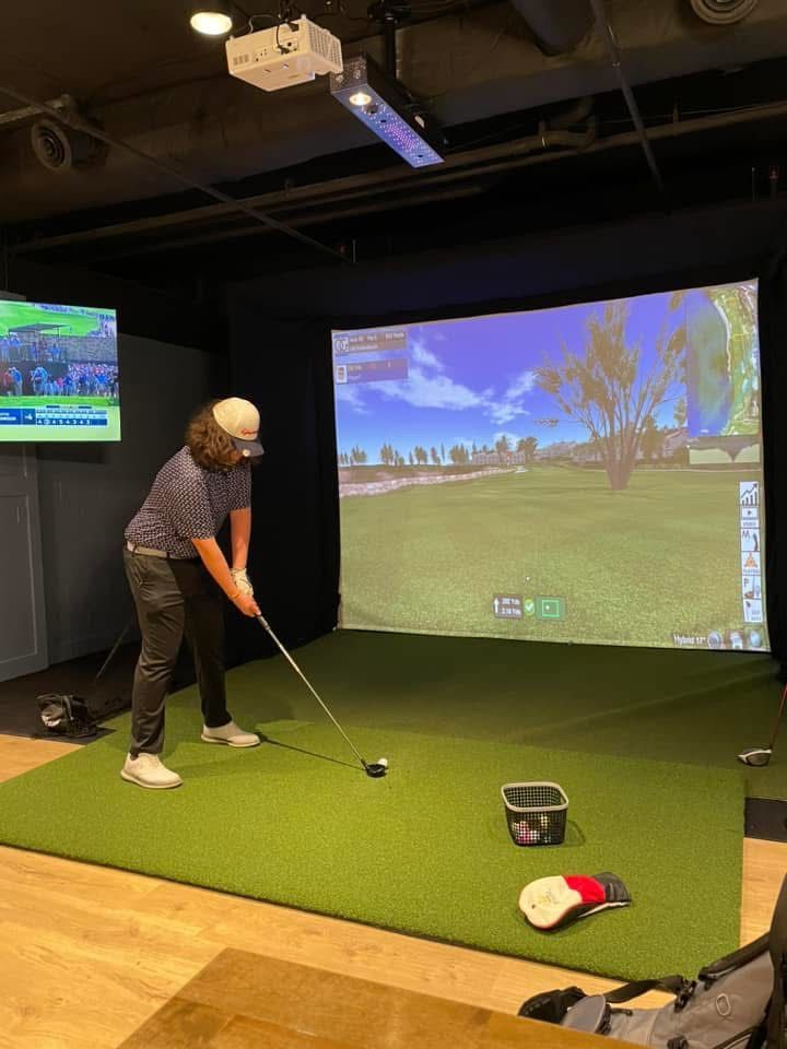 Person swings golf club in a golf simulator. Green turf, black background. Screen displays golf course.