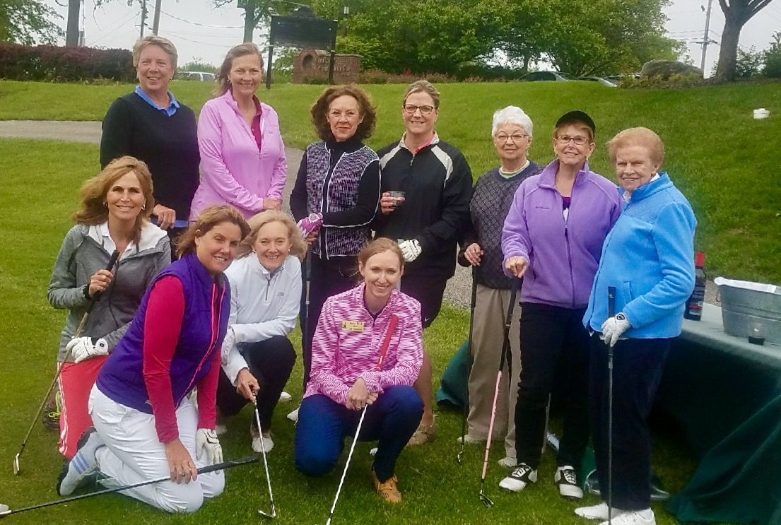 Group of women smiling on a golf course, holding golf clubs.