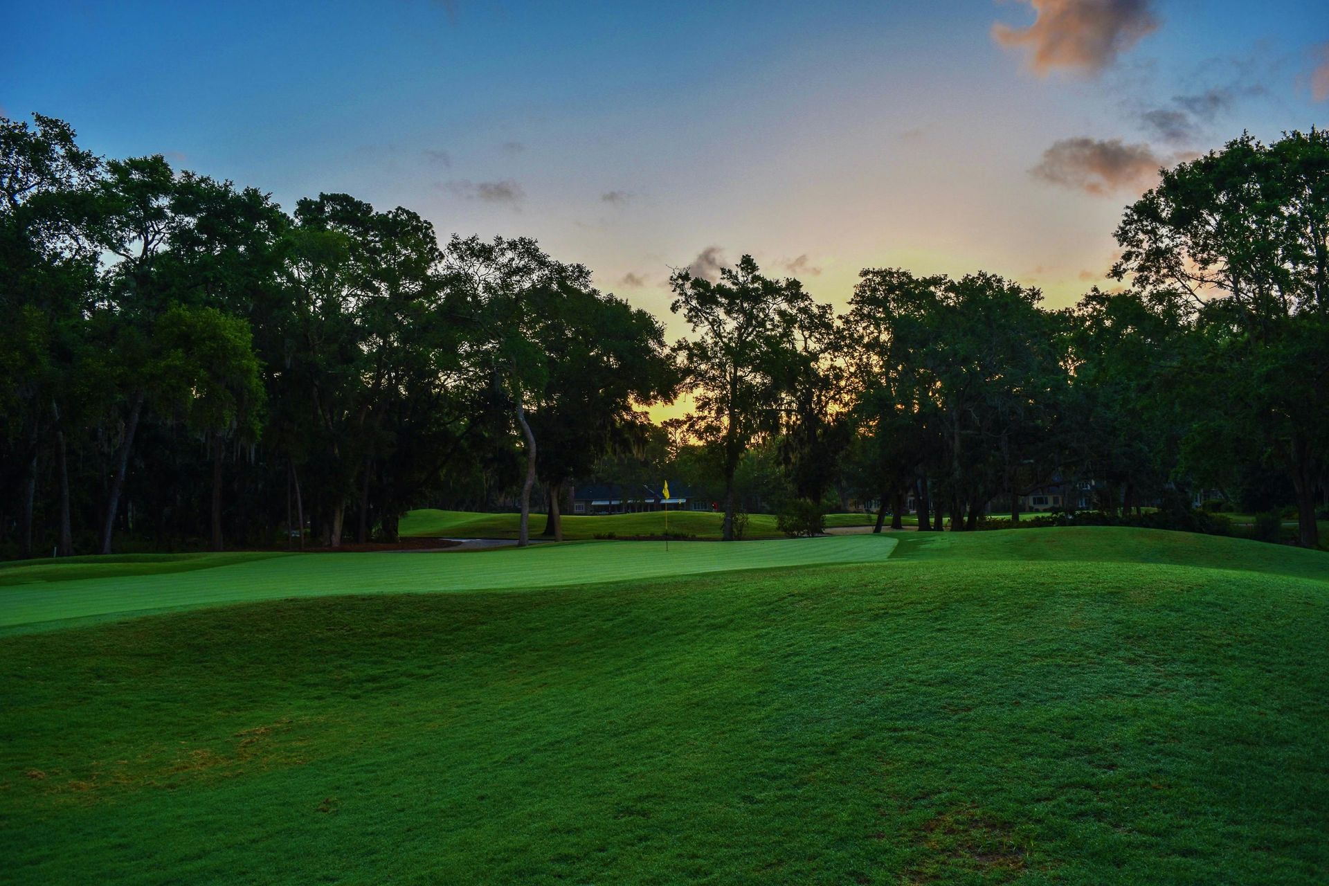 Green golf course at dusk with trees and a colorful sky.
