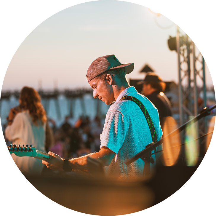 Musician playing guitar on an outdoor stage at sunset, with warm blurred lights in the background