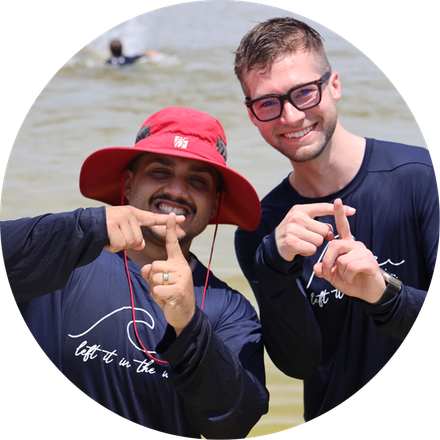 Two smiling friends in dark shirts and hats making finger-heart gestures at the beach