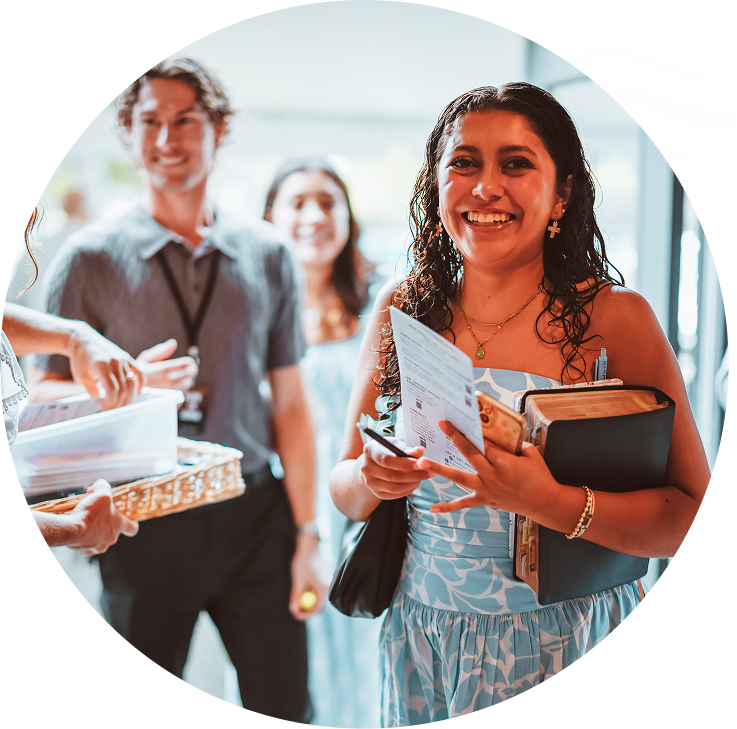 Smiling woman holding papers and a book, walking through a bright event venue with others behind her
