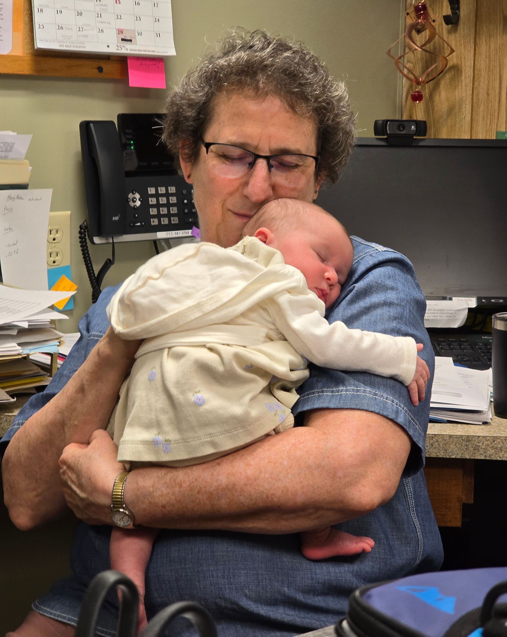 A person with curly hair and glasses gently holds a sleeping infant against their chest in an office setting.