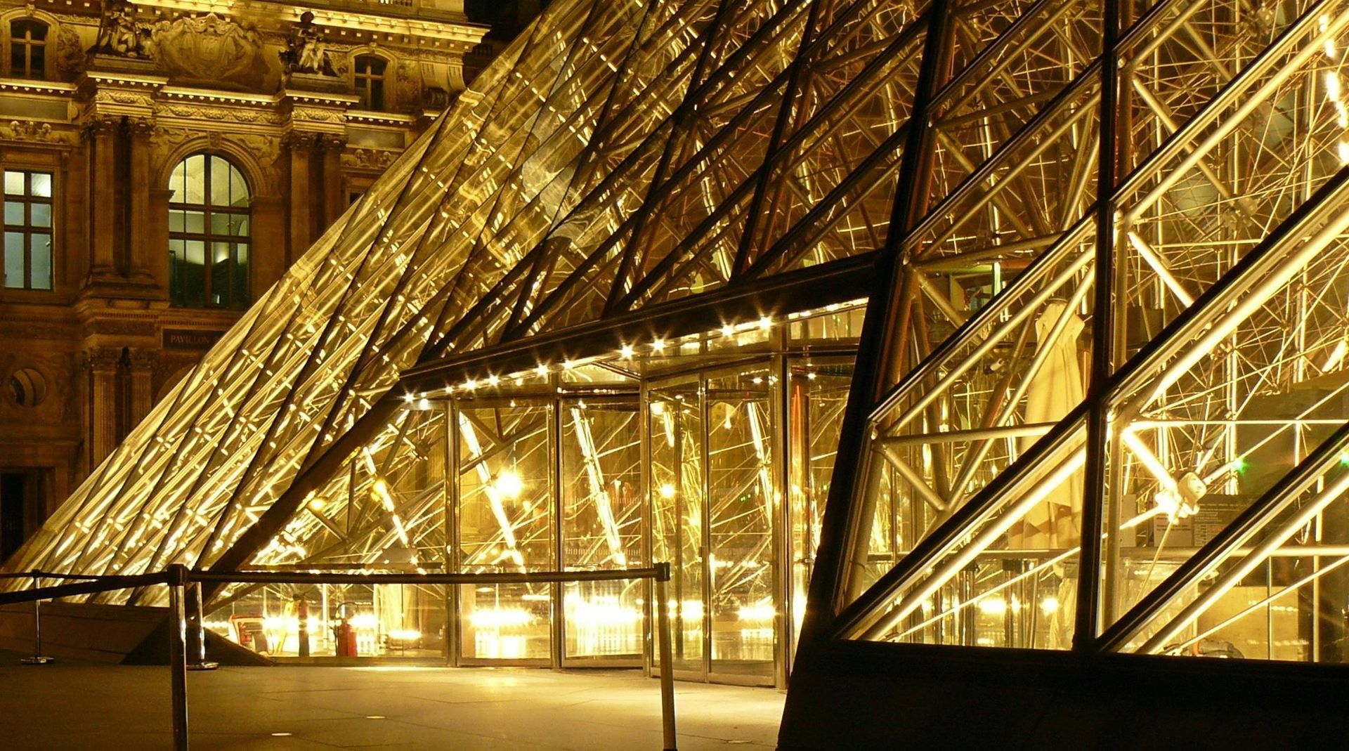 The Louvre Pyramid in Paris at night, illuminated with warm golden lights against a dark, classic stone building background.