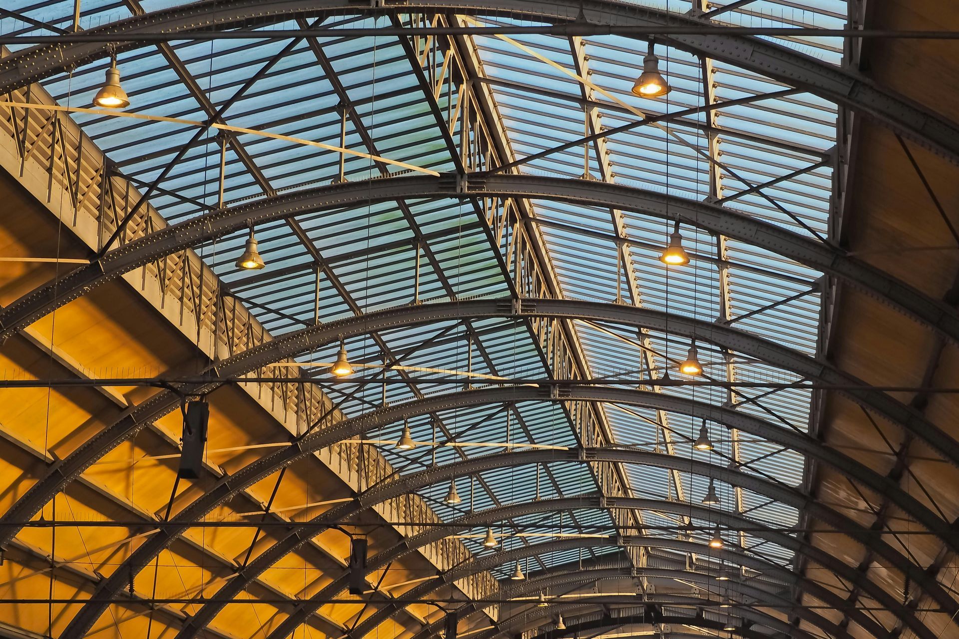 A vaulted glass-and-steel ceiling of a train station or conservatory, illuminated by hanging pendant lights.