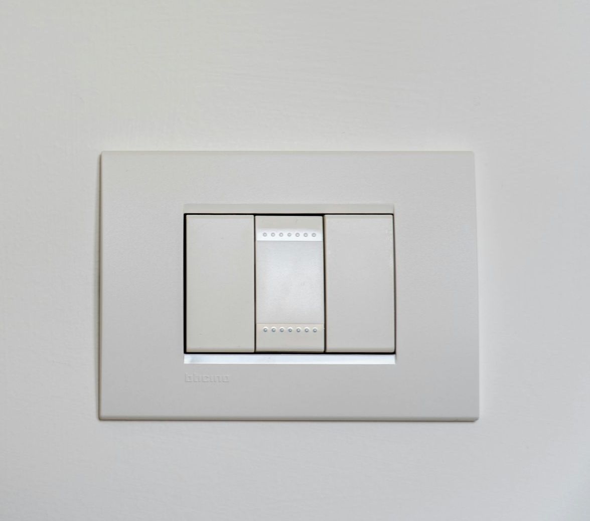 A white, rectangular triple light switch mounted on a white wall with a silver decorative trim in the center.