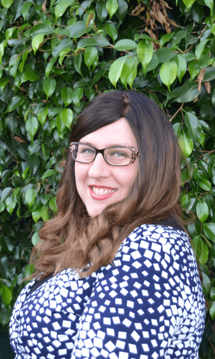 Smiling person with glasses in a blue patterned top standing in front of green foliage