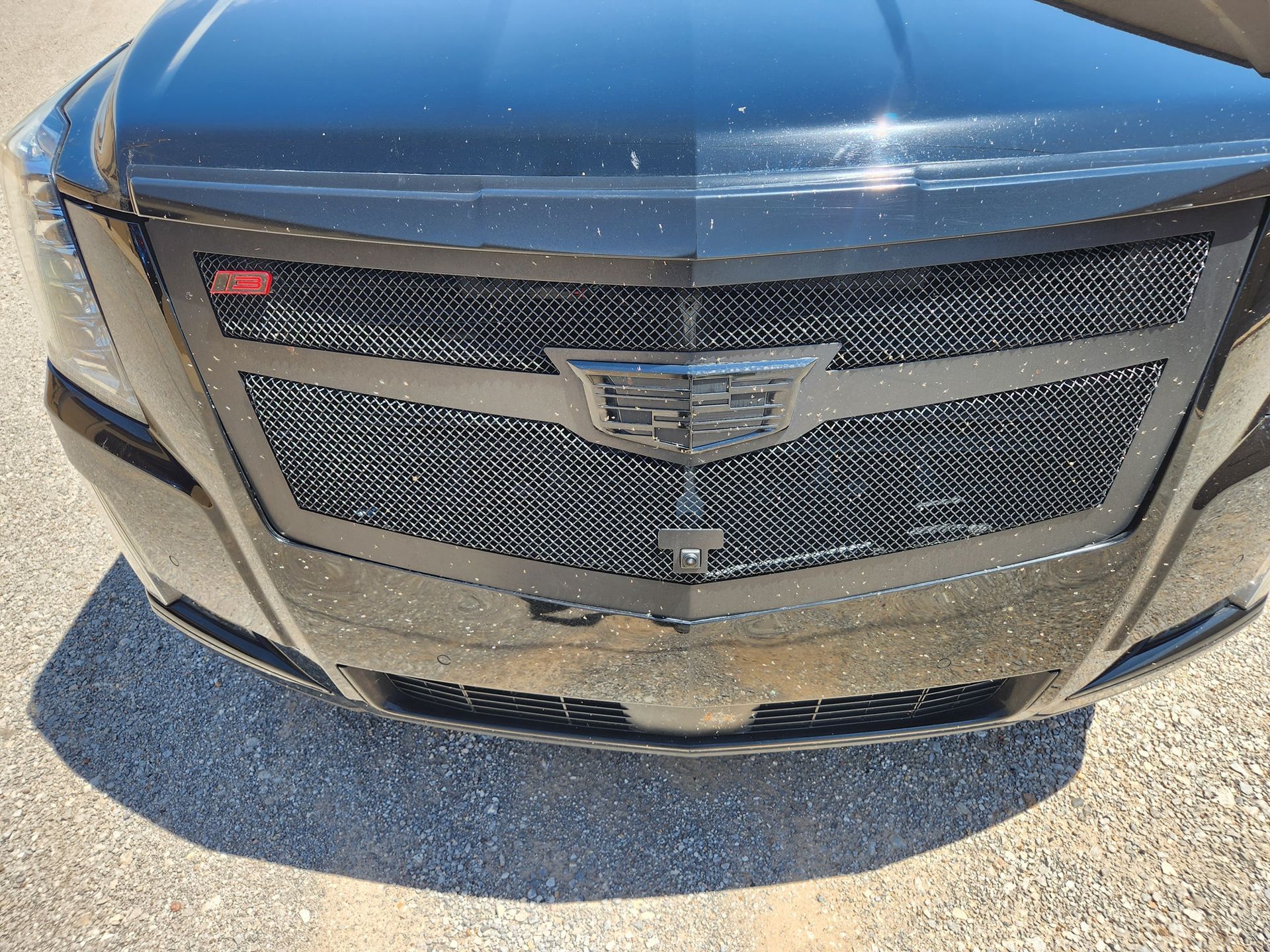 Close-up of a black Cadillac SUV front grille and emblem, with dusty bumper on pavement.