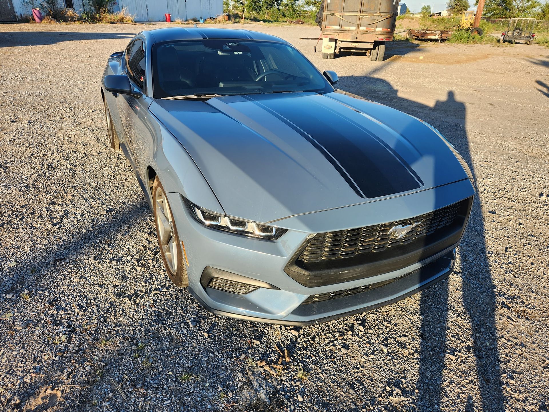 Silver sports car parked on gravel, viewed from the front-left in bright sunlight.