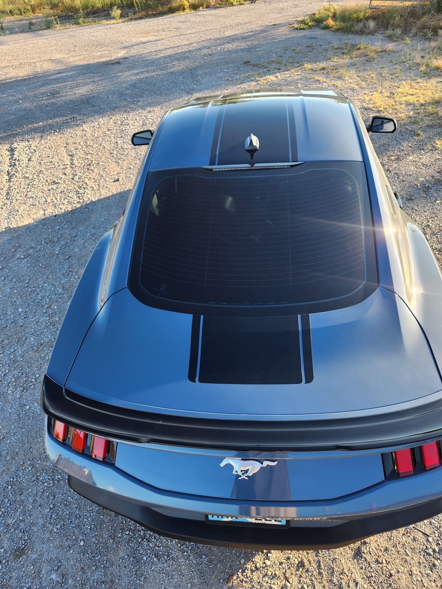 Blue Ford Mustang parked on gravel, viewed from above with a black roof and striped rear panel