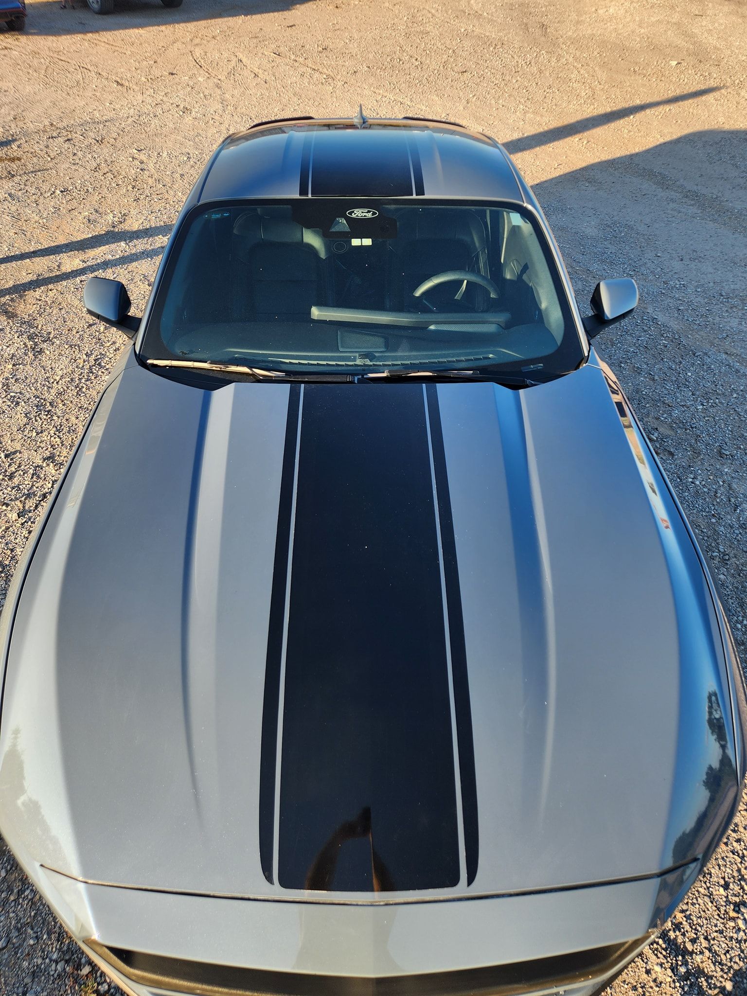 Silver sports car with black racing stripes, viewed from above on gravel.