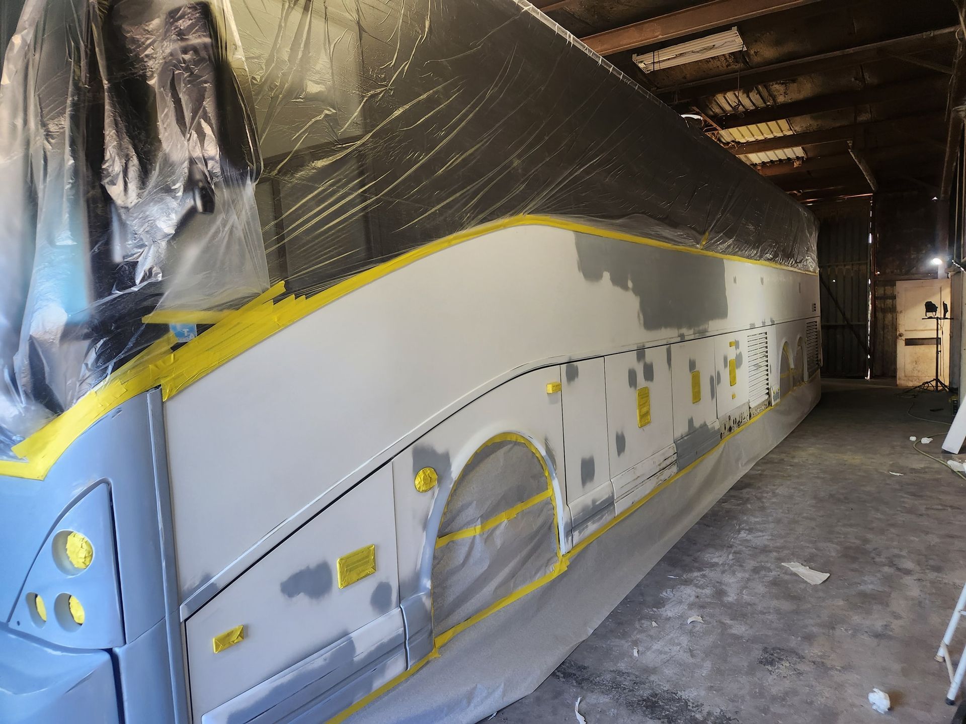 Partially painted bus body inside a workshop, covered in masking tape and plastic.