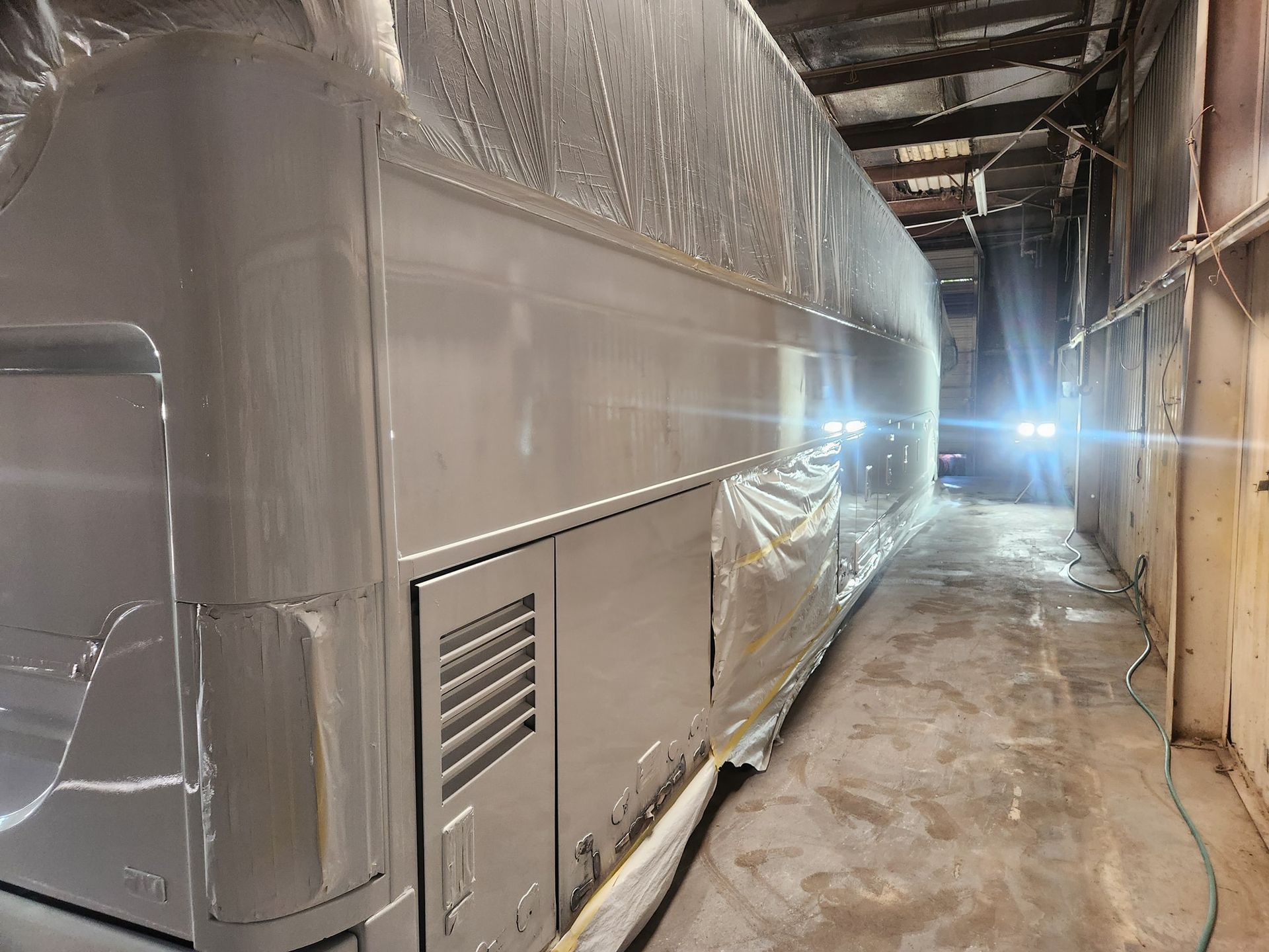 Large silver bus or trailer wrapped in plastic inside a dim industrial warehouse corridor.