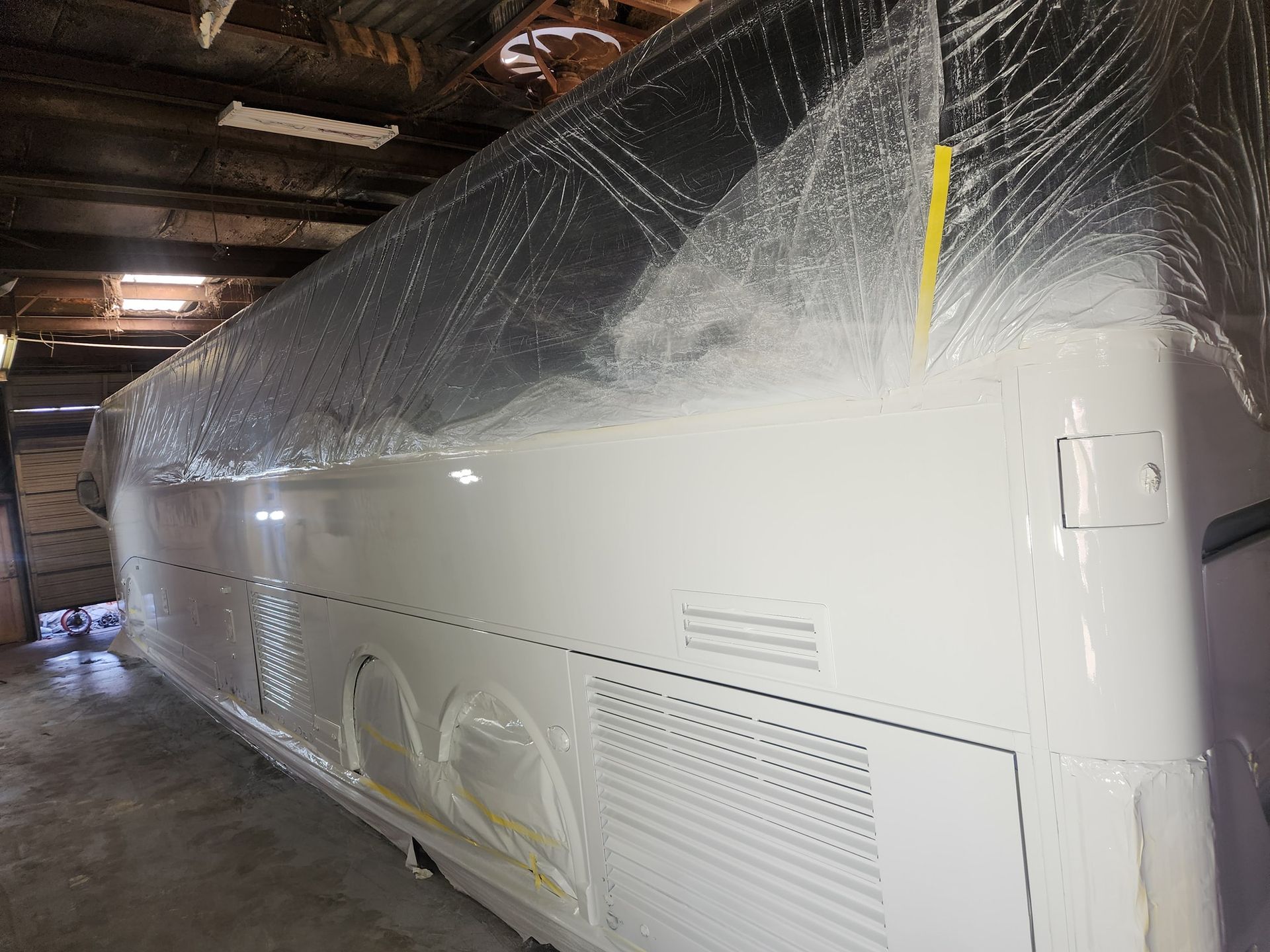 Large white bus wrapped in protective plastic inside a dim garage or warehouse.