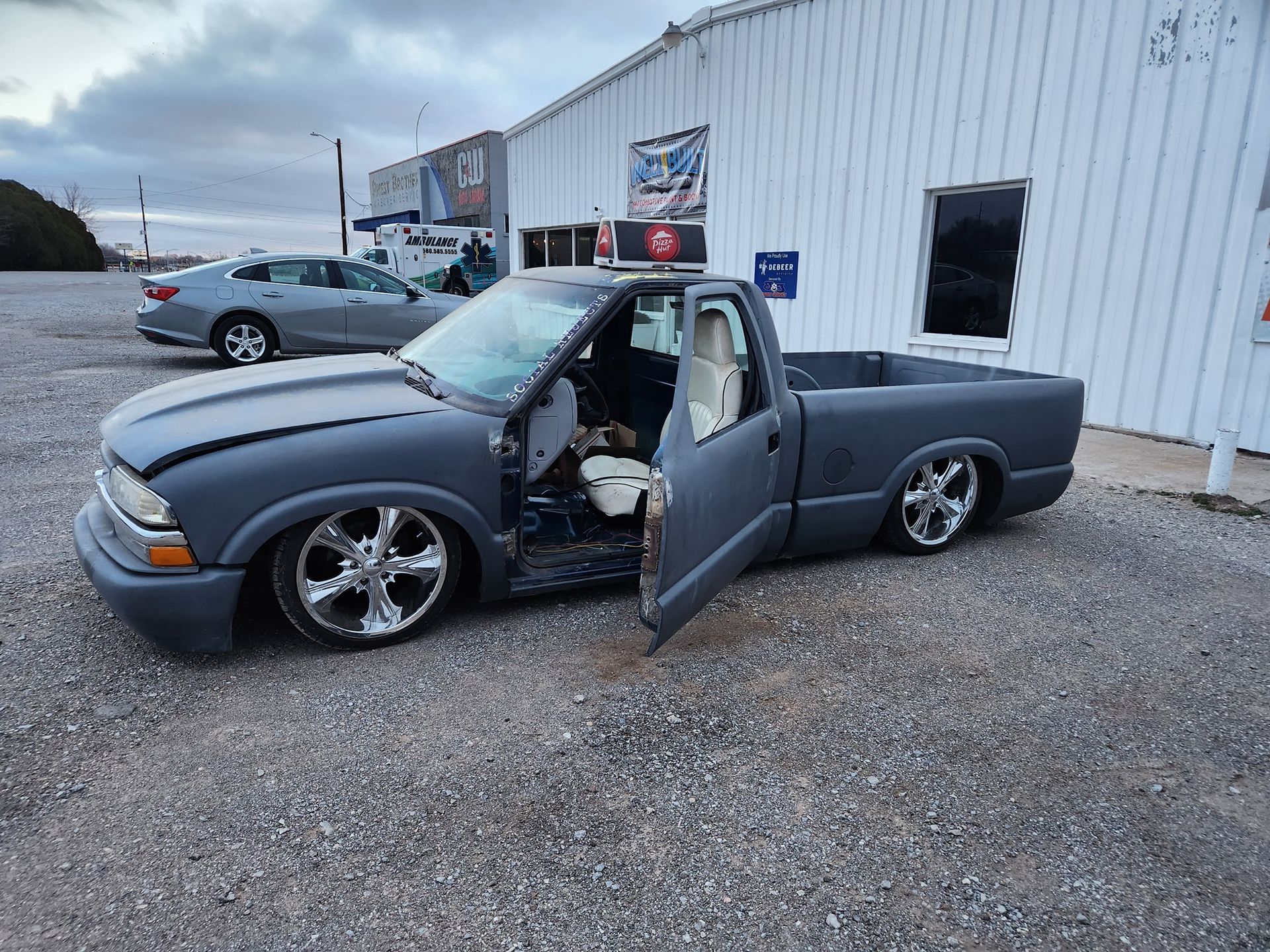 Lowered gray pickup truck with open door parked beside a white building on gravel.
